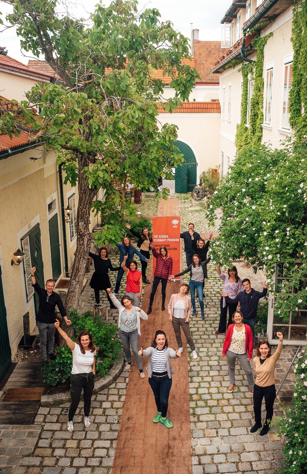 Group Picture of the program in Central and Eastern Europe in 2021, standing in a long-stretched courtyard with light houses and trees. Arms raised.