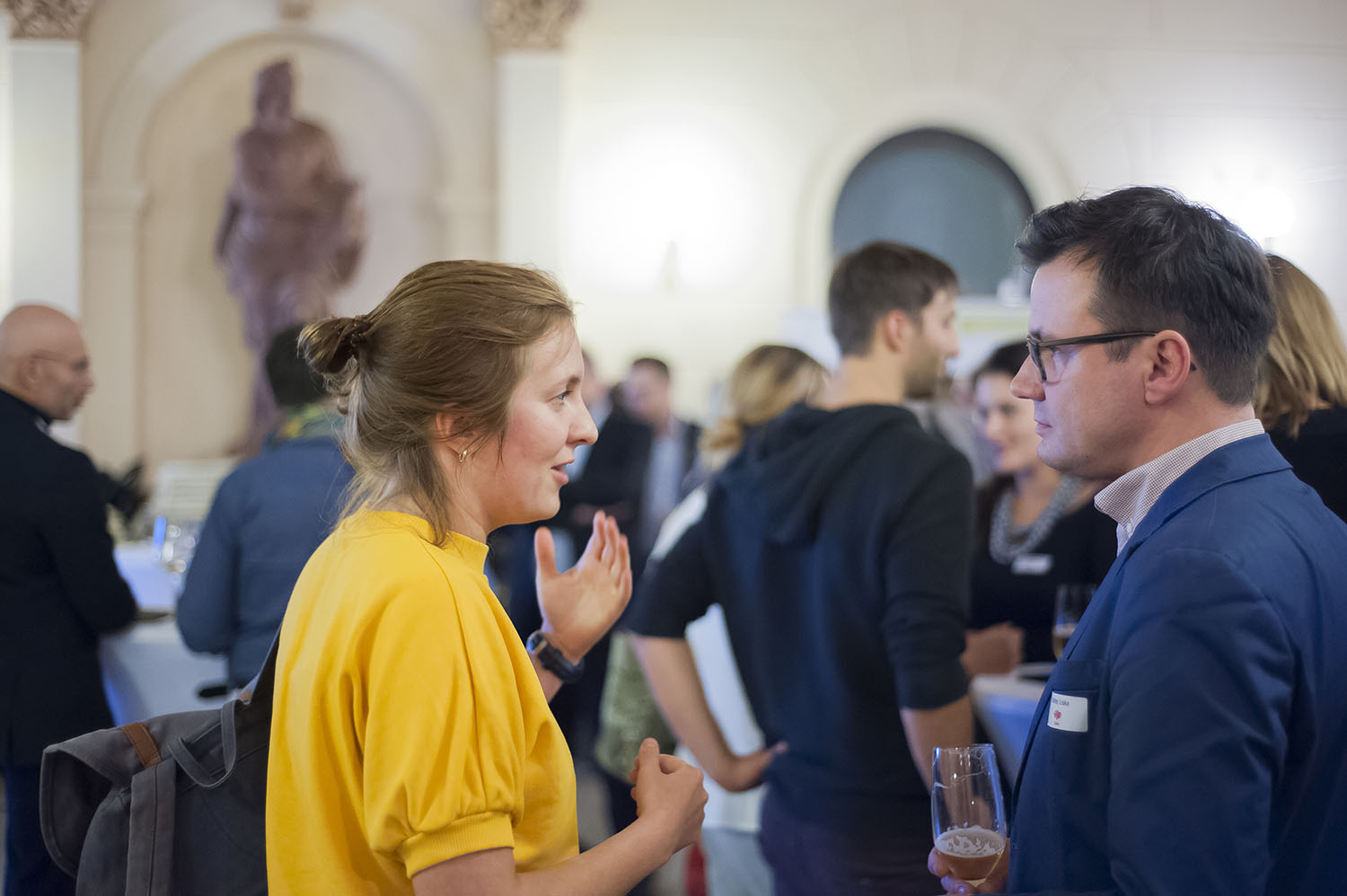 Networking. Young woman in a yellow shirt and man in a blue suit speaking at the Ashoka Visionary Program Kick-Off 2017, with more conversations in the background.