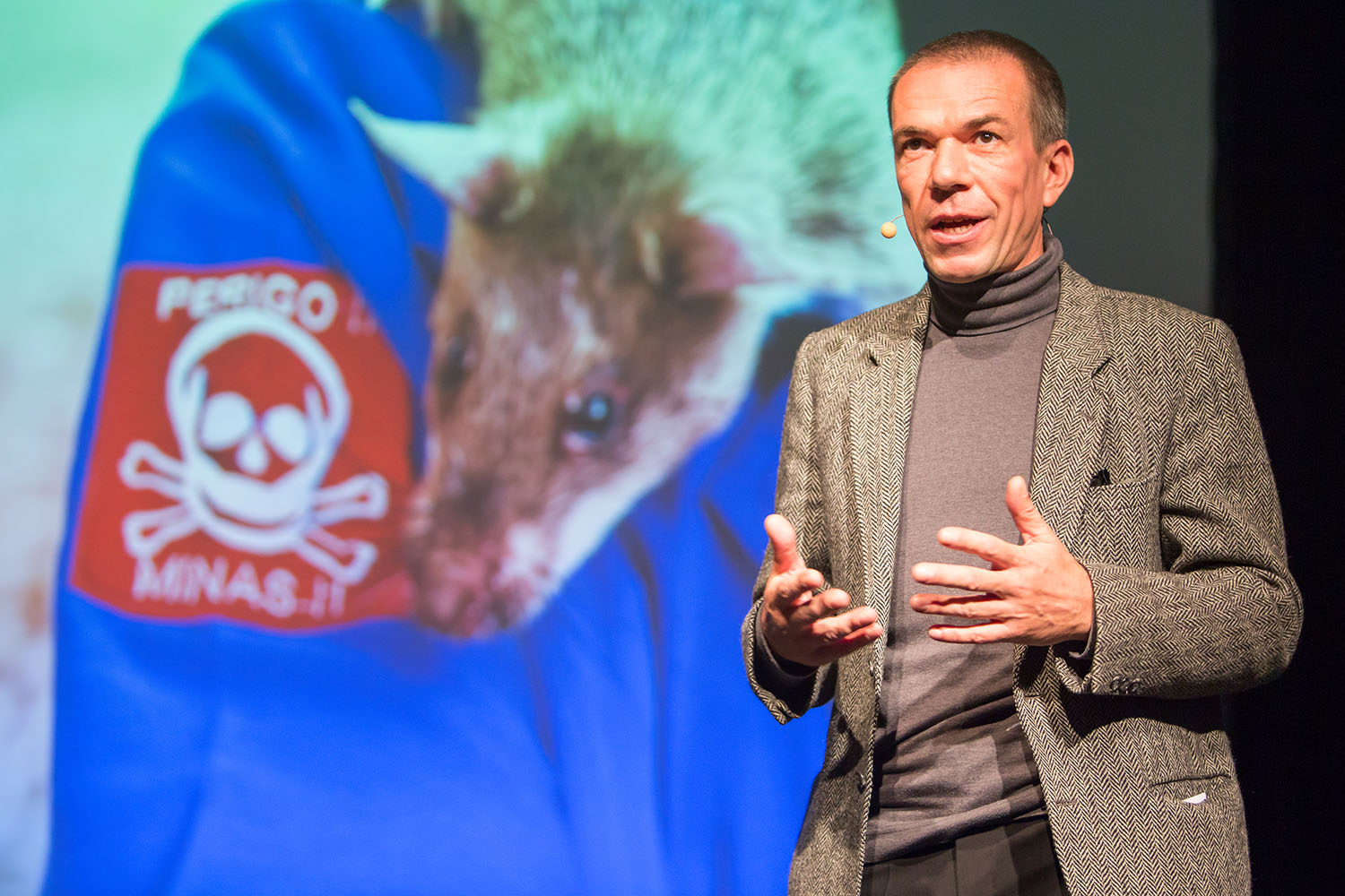 Ashoka Fellow Bart Weetjens is part of our network. Standing in a grey suit, he speaks from the scene of the Ashoka Visionary Program Launch in 2015. The screen in the background showes a a red badge reading "Caution! Mines!" and one of the HeroRats, who detect countless buried land mines.