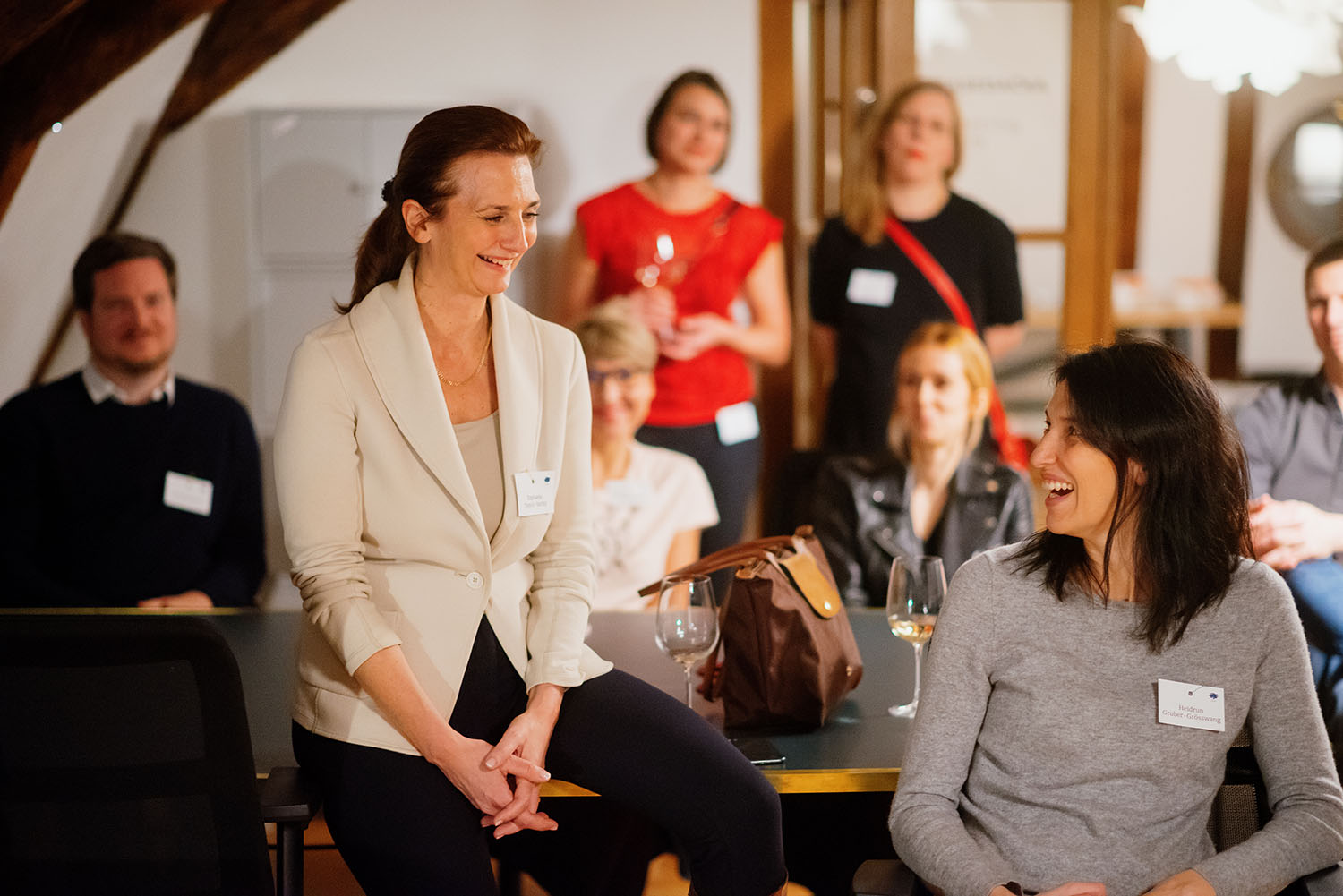 Evening session in the program cohort, with team spirit and inspiring insights. Image depicts two women sitting in the foreground and smiling at each other, and other people in the background of the room.