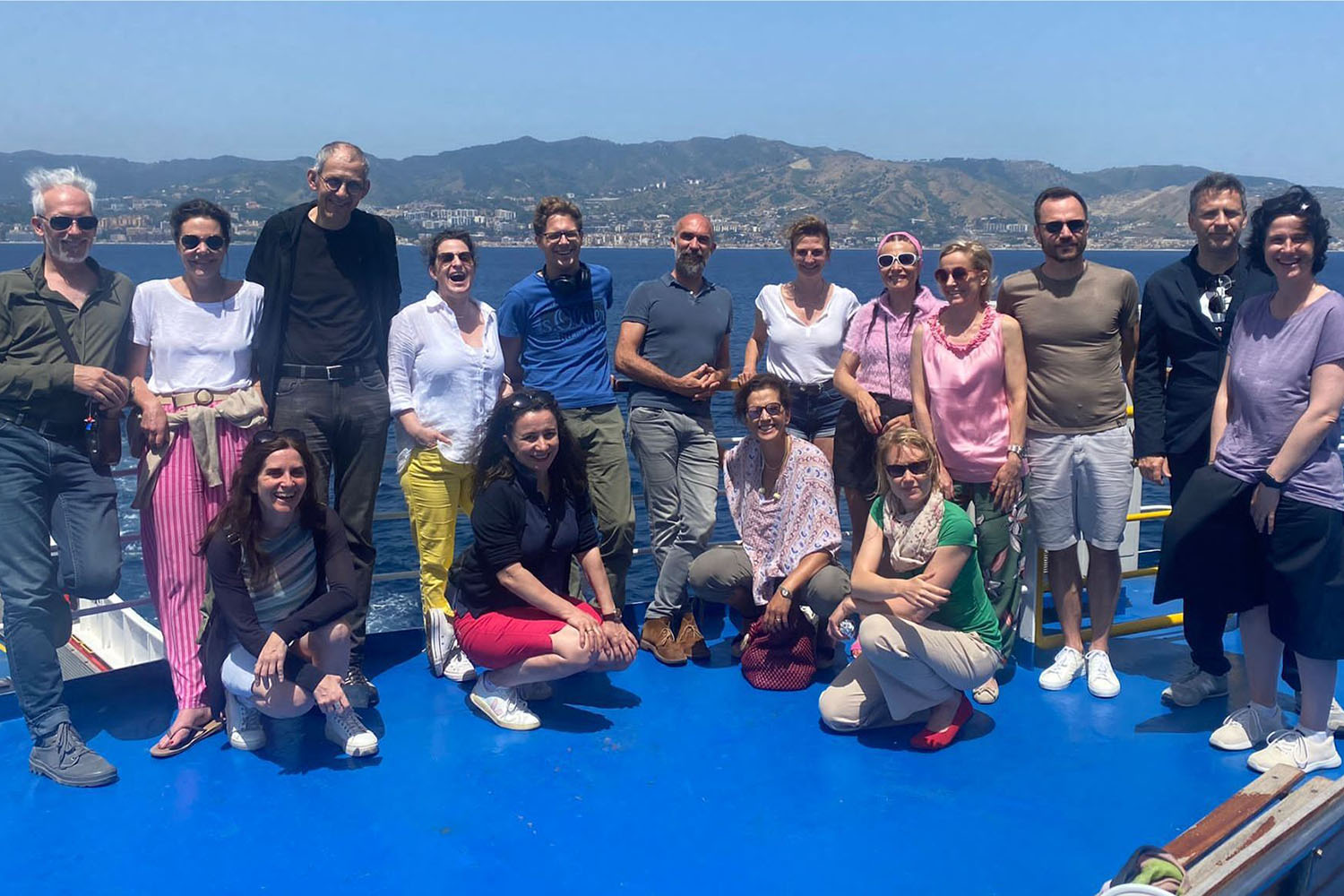 Alumni of the program join our network and benefit from journeys to changemakers. Participants of the Ashoka Visionary Journey to Calabria & Sicily, June '22, pose for a group photo on the blue deck of a ship, sea and island in the background.