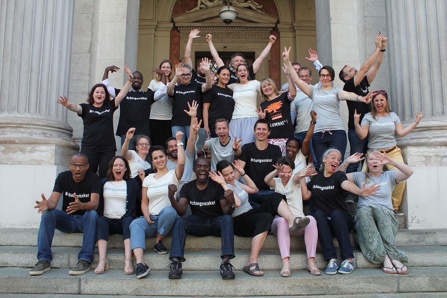 Group of Ashoka Visionary Program participants and alumni sitting on the steps to the Ashoka Austria Office Vienna, and raising their arms into the air full of energy.