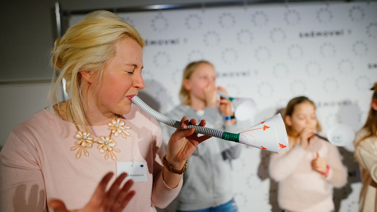 The program is playful, full of creativity and surprises. In the image, a participant of th Ashoka Visionary Program in Central and Eastern Europe, with blond hair and a rose short, blows into self-made paper trumpet. Along with children in the background.