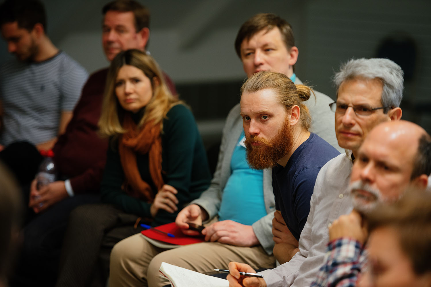 New learnings through each of the 7 modules of the program. In the close-up image, a bearded young man and other participants is sitting in the event room and listening intently to the speaker.